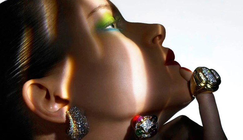 Close-up profile of a model with vibrant eye makeup, jeweled rings, and earrings against a white background