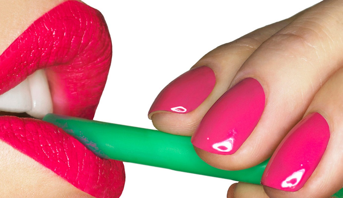 Close-up of a hand with pink nails holding a green object to bright red lips with visible white teeth against a white background.
