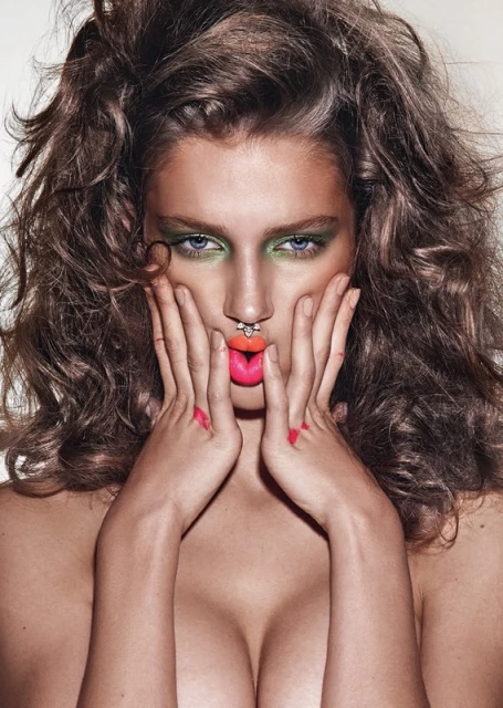 Close-up beauty shot of a young adult woman with wavy brown hair, green eyeshadow, a lip piercing, and bright pink lipstick.
