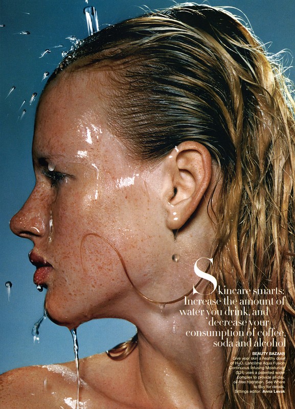 Close-up of a woman's face and upper body as water is poured on her. The photo shoot highlights skincare and beauty with wet skin and hair.