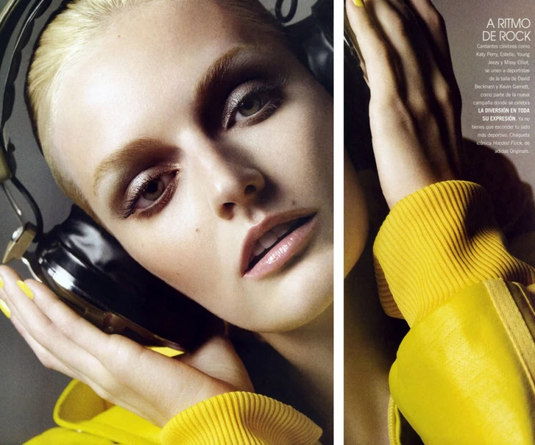 Close-up of a blonde woman wearing black headphones and a yellow jacket with a determined gaze, illuminated by soft, diffused lighting.
