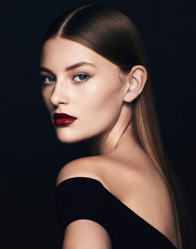 Portrait of an adult woman with dark red lipstick, wearing a black off-the-shoulder top, posing against a dark background in a photo studio.