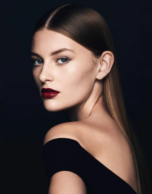 Portrait of an adult woman with dark red lipstick, wearing a black off-the-shoulder top, posing against a dark background in a photo studio.