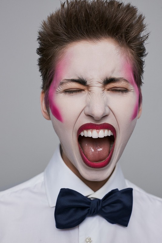 Close-up of a white-skinned adult with pink makeup and a dark bow tie, face contorted in a scream, mouth open wide showing teeth.