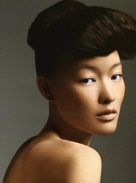 Portrait of a woman with brown hair styled up, gazing over her shoulder in a beauty shot against a grey backdrop with even lighting.