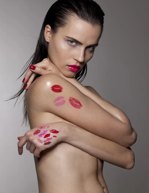 A nude female model poses with vibrant lipstick kiss marks on her arm and hand against a neutral grey background in a beauty shot.
