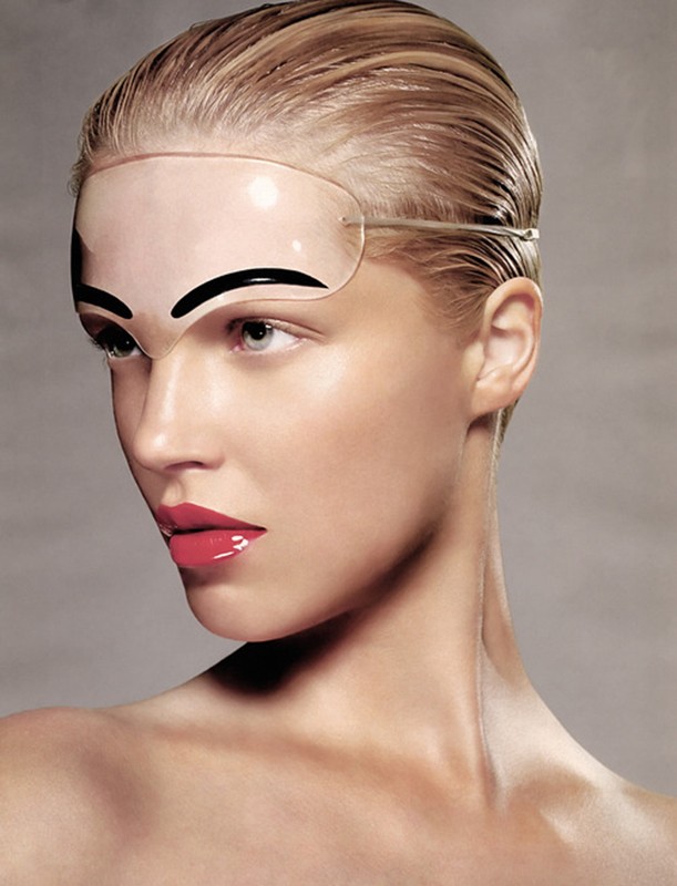 Woman with glossy makeup and slicked-back hair poses in a close-up studio shot, wearing a unique clear plastic eye shield with painted eyebrows.