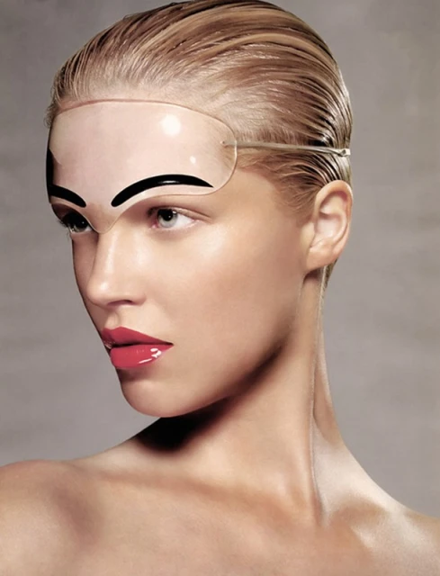 Woman with glossy makeup and slicked-back hair poses in a close-up studio shot, wearing a unique clear plastic eye shield with painted eyebrows.