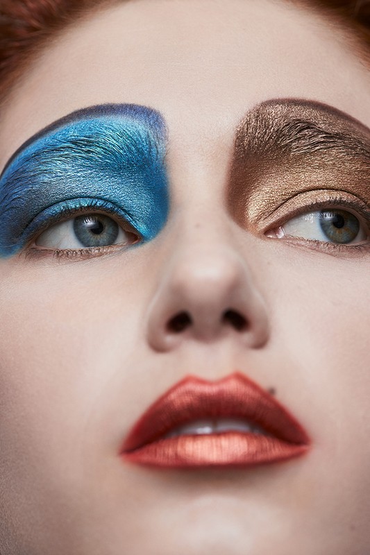 Close-up portrait of a woman with striking makeup: blue eyeshadow on one eye, bronze on the other, and metallic red lips.
