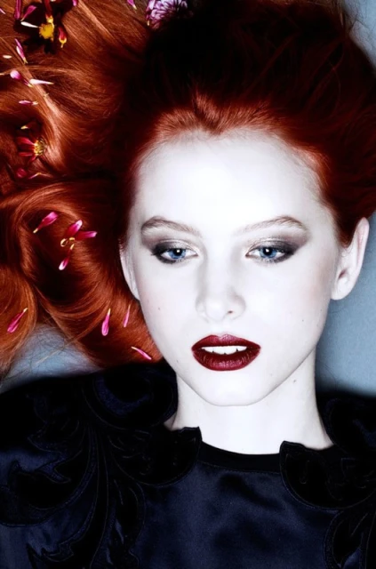 Close-up beauty shot of a fair-skinned adult female model with vibrant red hair adorned with delicate flowers, dark eyeshadow and lipstick.