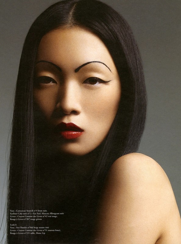 Close-up portrait of a woman with fair skin, dark hair, red lips, and stylized eyebrows, posing against a gray backdrop in studio lighting.