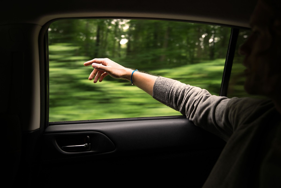 Arm extended from a car window; blurred landscape with green foliage in the background, illuminated by natural light.