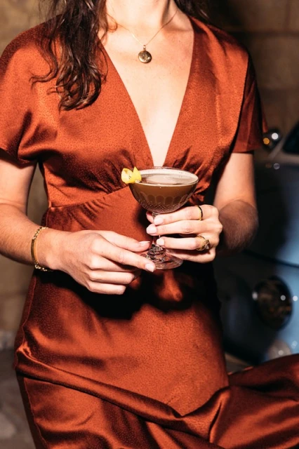 Woman in an orange satin dress holds a cocktail in a coupe glass with a lemon twist garnish, posing next to a vintage car.