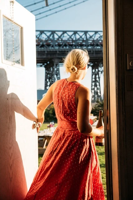 Woman in red polka dot dress exiting white door onto green lawn with wine, bridge in background, bathed in warm sunlight.