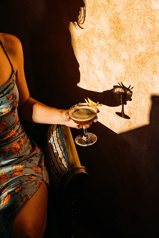 Woman in floral dress holds a cocktail with lemon twist. Shadow of a person also holding a cocktail in the background.