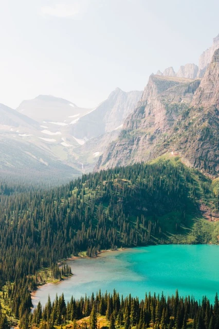 Overhead view of a bright turquoise mountain lake surrounded by a dense evergreen forest and rugged mountain peaks on a clear day.