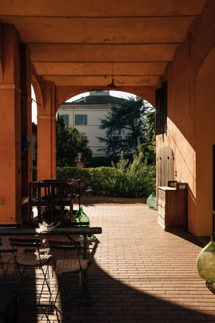 Outdoor patio with furniture, lush greenery, and a building under a covered archway, bathed in warm sunlight creating strong shadows.