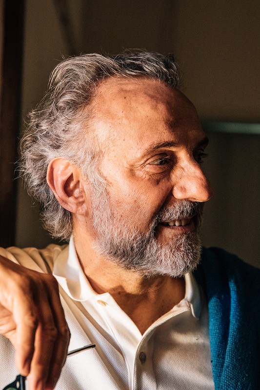 Close-up portrait of a senior adult man with gray hair and beard. He is indoors with natural light, wearing a white shirt and sweater.