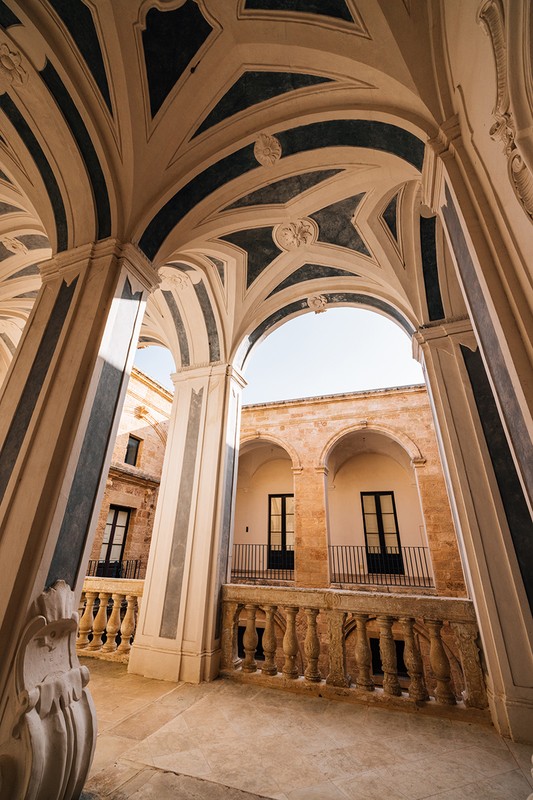 Architectural photography of a stone balcony with arches and columns, under a decorated ceiling with blue stripes and designs.
