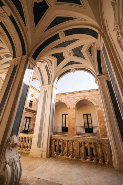 Architectural photography of a stone balcony with arches and columns, under a decorated ceiling with blue stripes and designs.