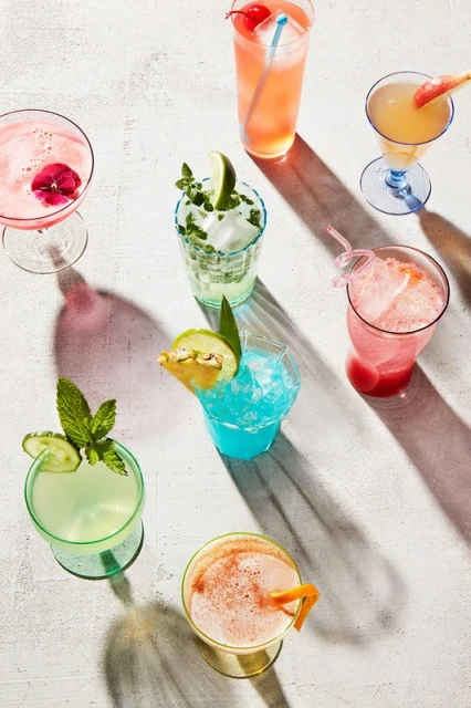 Overhead shot of colorful cocktails in assorted glasses, garnished with fruit and herbs, arranged on a textured white surface in bright light.