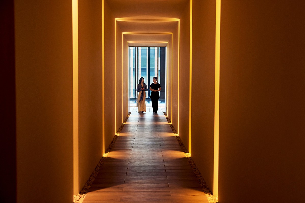Two women walking down a hotel hallway with illuminated recesses, bathed in warm light, and leading to a window with a city view.