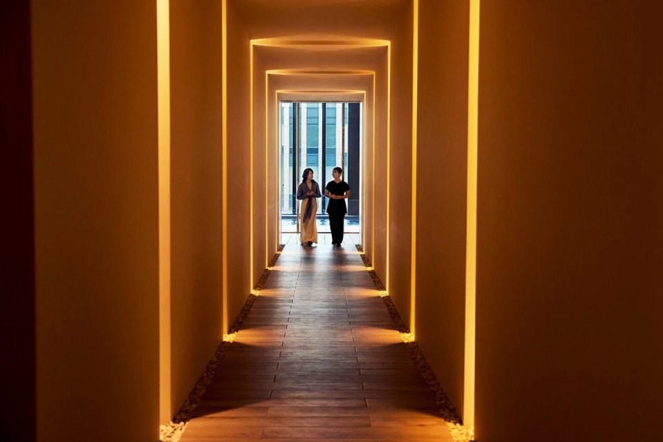 Two women walking down a hotel hallway with illuminated recesses, bathed in warm light, and leading to a window with a city view.