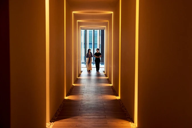 Two women walking down a hotel hallway with illuminated recesses, bathed in warm light, and leading to a window with a city view.