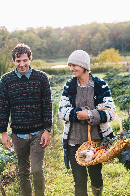 A happy couple walks through a sunny rural garden with fresh vegetables in a woven basket, wearing sweaters, and smiling.