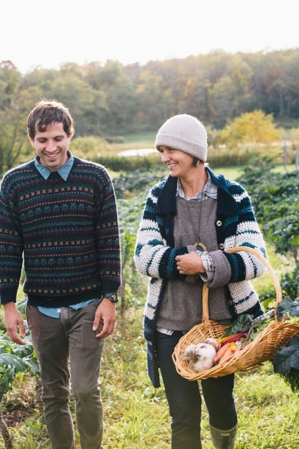 A happy couple walks through a sunny rural garden with fresh vegetables in a woven basket, wearing sweaters, and smiling.