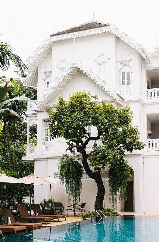 Exterior of a white hotel building by a pool with lounge chairs and a tree with weeping branches on a cloudy day.