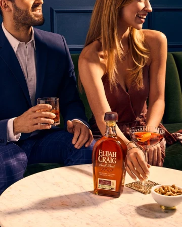 Elijah Craig whiskey bottle on a marble table with a man and a woman, cocktails, and snacks in a dimly lit lounge setting.