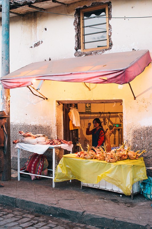 Outdoor view of a woman working at a food stand under an awning, with raw chickens and meat visible, and a window above the entrance.
