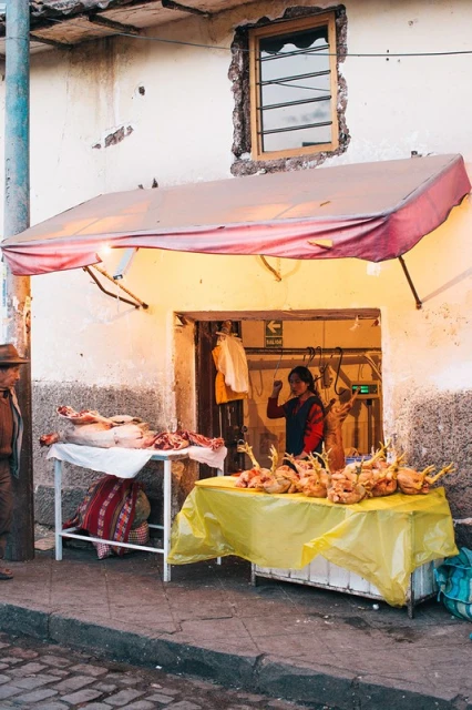 Outdoor view of a woman working at a food stand under an awning, with raw chickens and meat visible, and a window above the entrance.