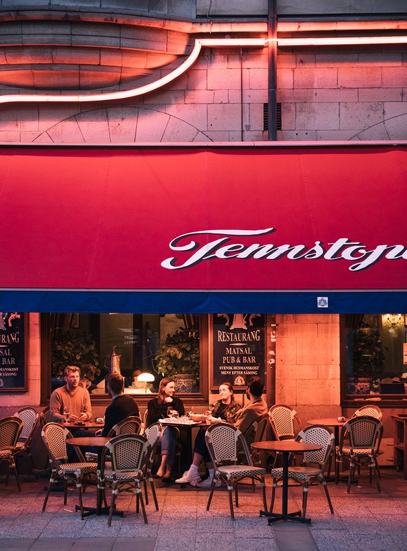 Outdoor restaurant scene with group of people seated at tables, enjoying food and drinks under a red awning with neon lighting.