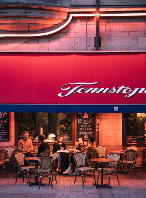 Outdoor restaurant scene with group of people seated at tables, enjoying food and drinks under a red awning with neon lighting.