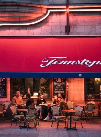 Outdoor restaurant scene with group of people seated at tables, enjoying food and drinks under a red awning with neon lighting.