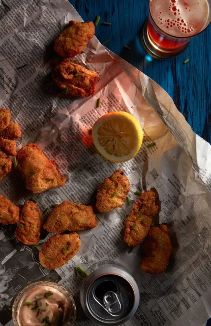 Overhead shot of fried food, lemon, and beer on newspaper and blue wood, showcasing food photography for culinary and lifestyle content.