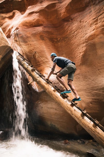 Man climbs log ladder next to waterfall in slot canyon with red rock walls. Adventurous travel and outdoor recreation theme.