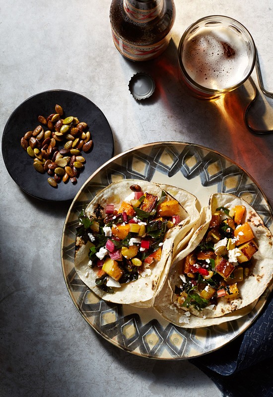 Overhead shot of two tacos on a patterned plate next to a small plate of seeds, a beer bottle, and a glass of beer.