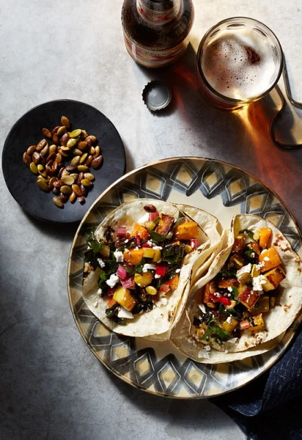 Overhead shot of two tacos on a patterned plate next to a small plate of seeds, a beer bottle, and a glass of beer.