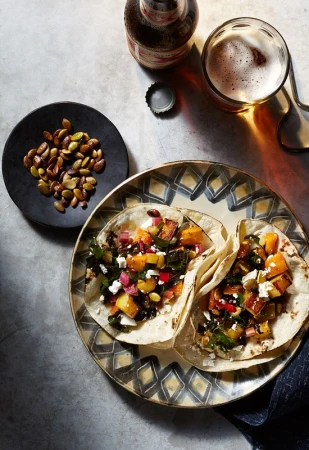 Overhead shot of two tacos on a patterned plate next to a small plate of seeds, a beer bottle, and a glass of beer.