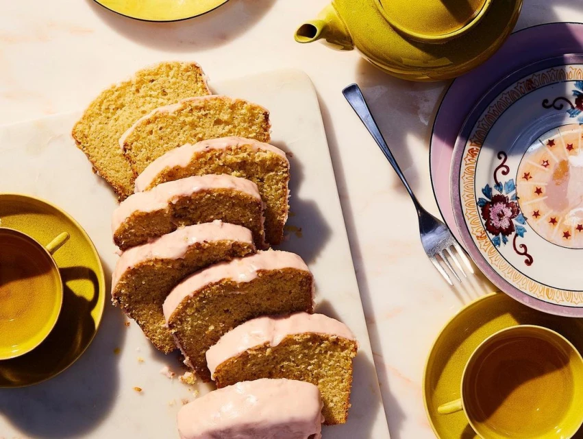 Overhead shot of sliced pound cake with pink icing, served on a marble board with yellow teacups and a yellow teapot.