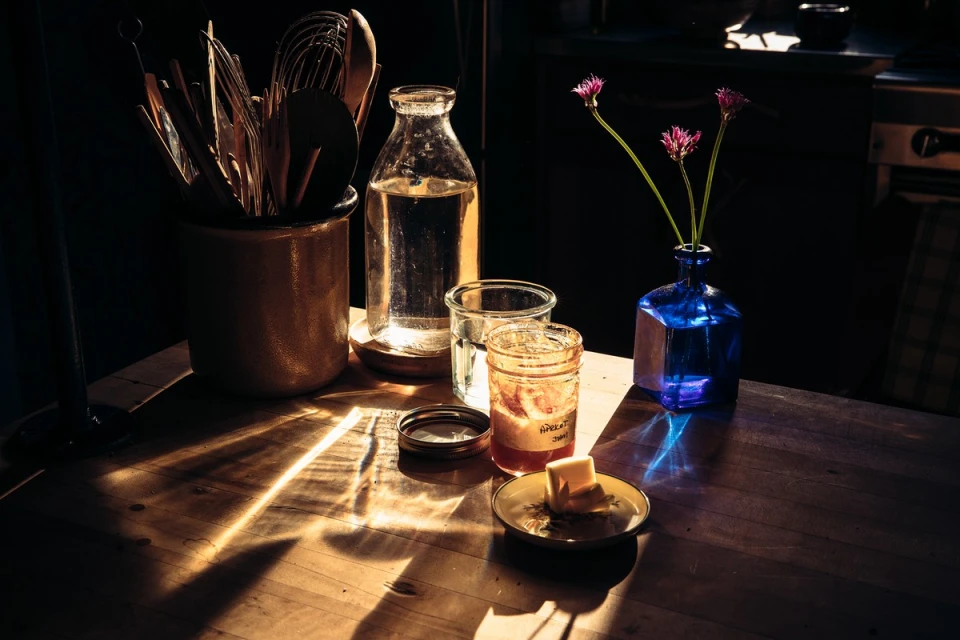 Still life photograph of a rustic wooden table with cooking utensils, jars, a blue vase with flowers, and reflections of light.