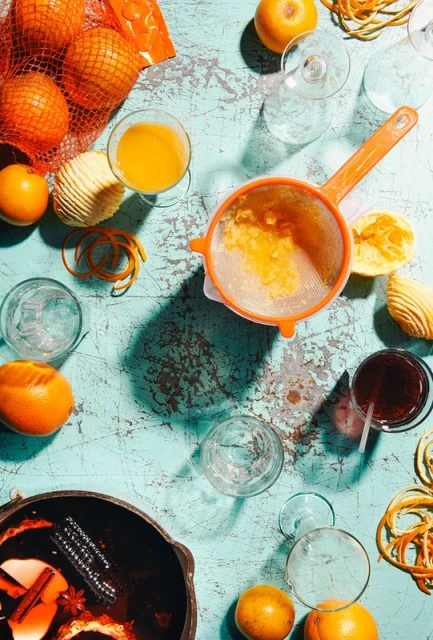 Overhead shot of orange juice and oranges in a mesh bag next to a pot of mulled wine on a distressed aqua surface with scattered glassware.