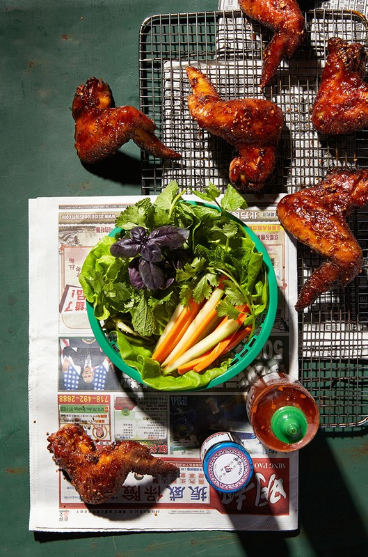 Overhead shot of cooked chicken wings on a cooling rack, accompanied by a basket of fresh herbs and vegetables, and two condiment bottles.