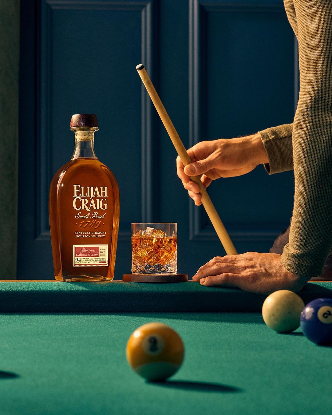 Elijah Craig bourbon whiskey bottle and a rocks glass on a pool table with a pool cue held by a man wearing a tan sweater.