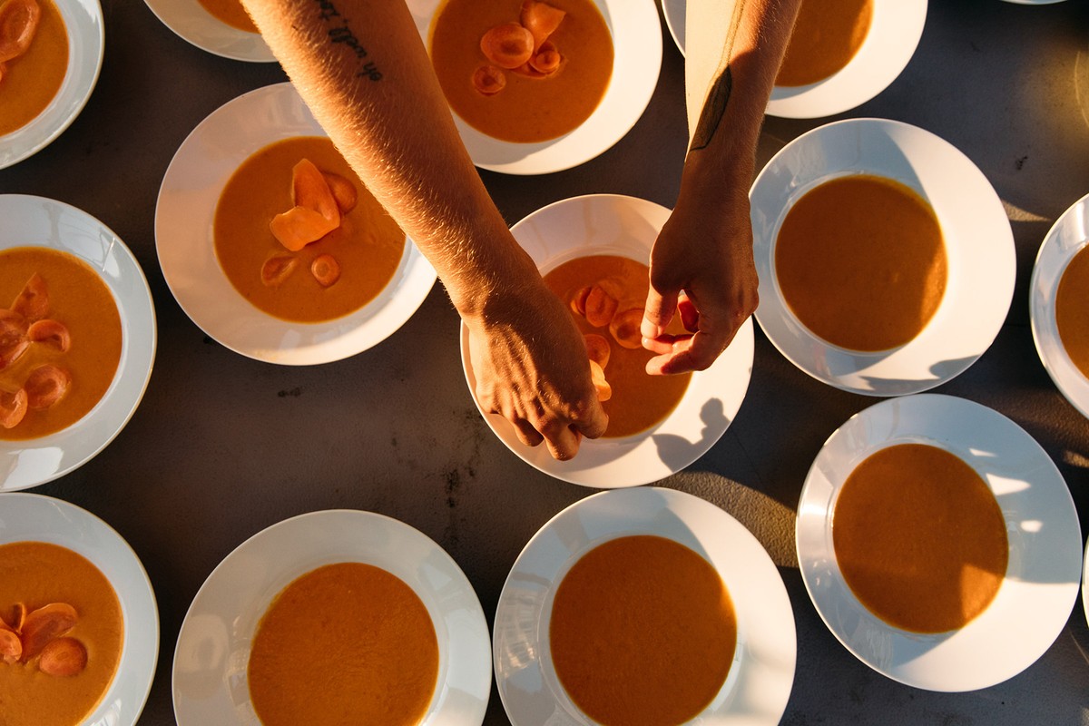 Overhead view of a row of white bowls filled with orange soup being prepared by a person with tattooed arms.