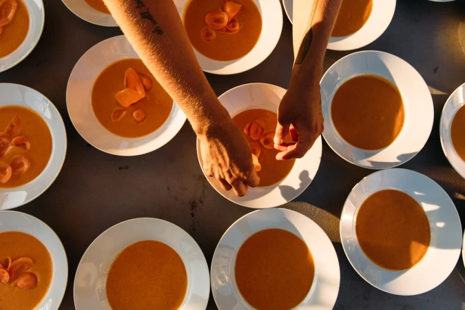 Overhead view of a row of white bowls filled with orange soup being prepared by a person with tattooed arms.