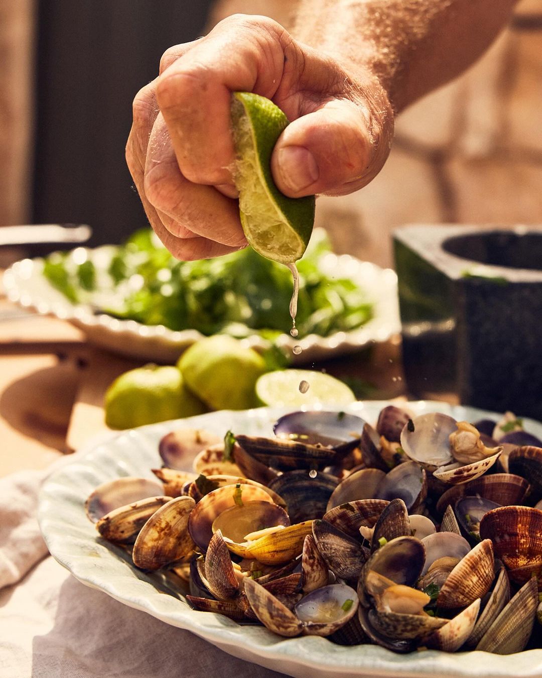 Lime juice is squeezed over clams in a white bowl with more limes and greens in the background on a wood surface. Food photography.